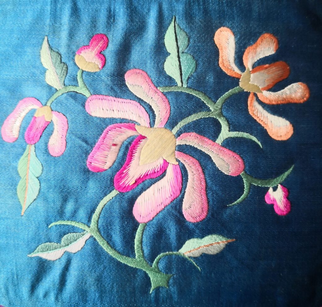Pink and peach floral embroidery with green stems on blue fabric, showing the direction and texture of the stitches