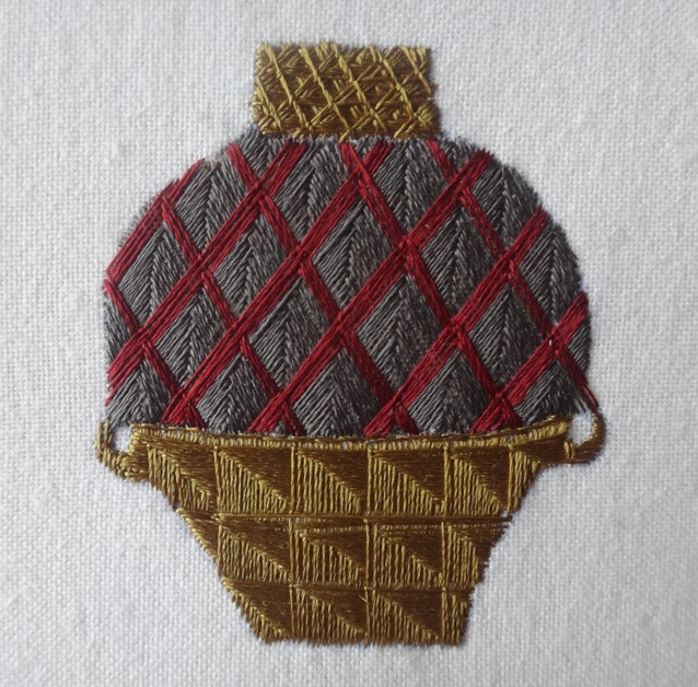 Embroidered jar on white cloth, with dark red diagonal lattice over a grey body and brown geometric patterns in its upper and lower sections.